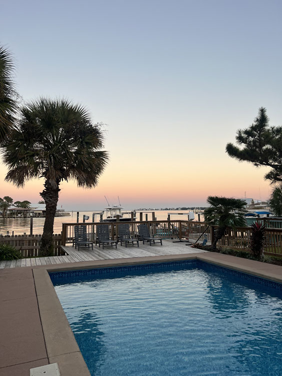 Sunset view of a pool deck by the waterfront with palm trees and boats in the distance